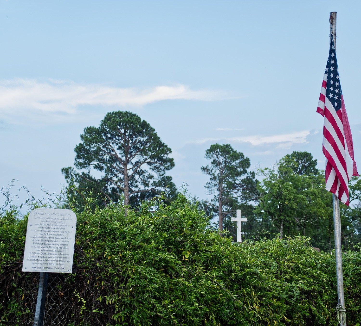 A historical marker and American flag stand just inside the fence between Cedar Memorial Gardens and the Black cemetery in Mineola, with a cross in the background on the other side. The fence is scheduled to be taken down next week.